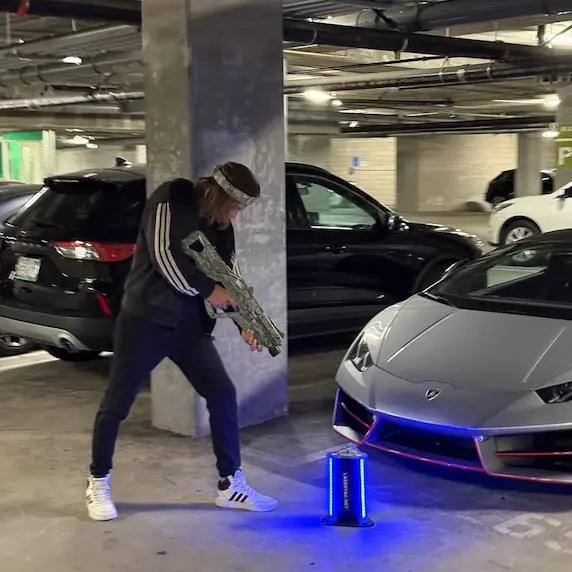 Man playing laser tag in an underground parking garage near a Lamborghini, aiming a laser tag rifle at a glowing blue target.