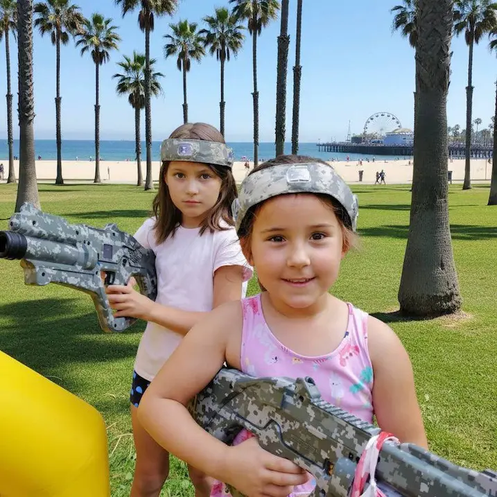 Two girls with laser tag blasters at Santa Monica beach with palm trees and the ocean in the background
