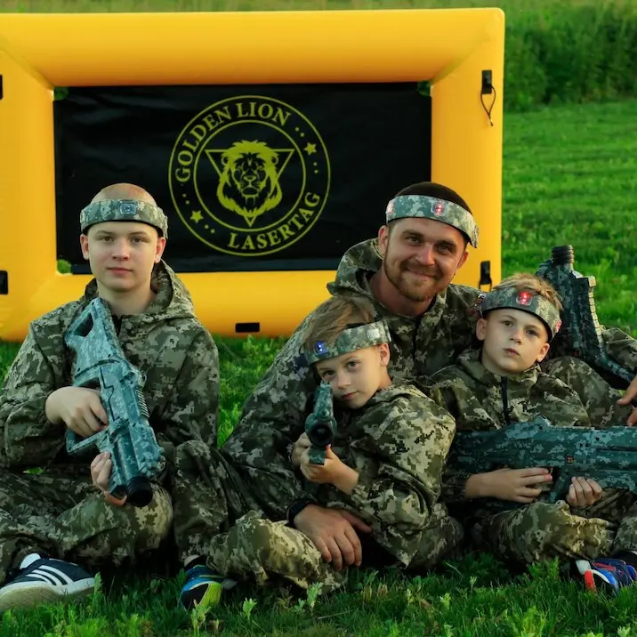 Group of kids in camouflage with an adult posing with laser tag blasters in front of a yellow inflatable barrier.
