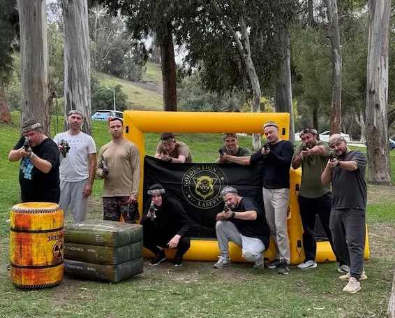 Group of men posing with laser tag guns in front of a Golden Lion Lasertag inflatable barricade at an outdoor park event.